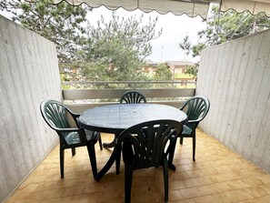 Eine überdachte Terrasse mit rundem Tisch und vier Stühlen, umgeben von Holzwänden und Blick auf Bäume und Gebäude.
