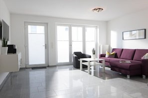 A modern living room with a sofa, a coffee table, and a chair. There is a sliding glass door leading to a balcony. - Seilerstr./Knyphausenstr.26A - Fiene (Norderney)