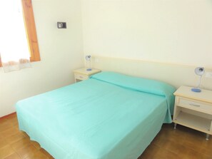 Een eenpersoonskamer met een blauwgroene sprei, twee nachtkastjes met lampen, een houten vloer en een raam met gordijn.