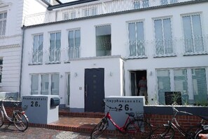 Ein weißes, zweistöckiges Wohnhaus mit einem Balkon im oberen Stockwerk, ein rotes Fahrrad davor geparkt und ein Briefkasten mit der Aufschrift 'Knyphausenstr. 26'.