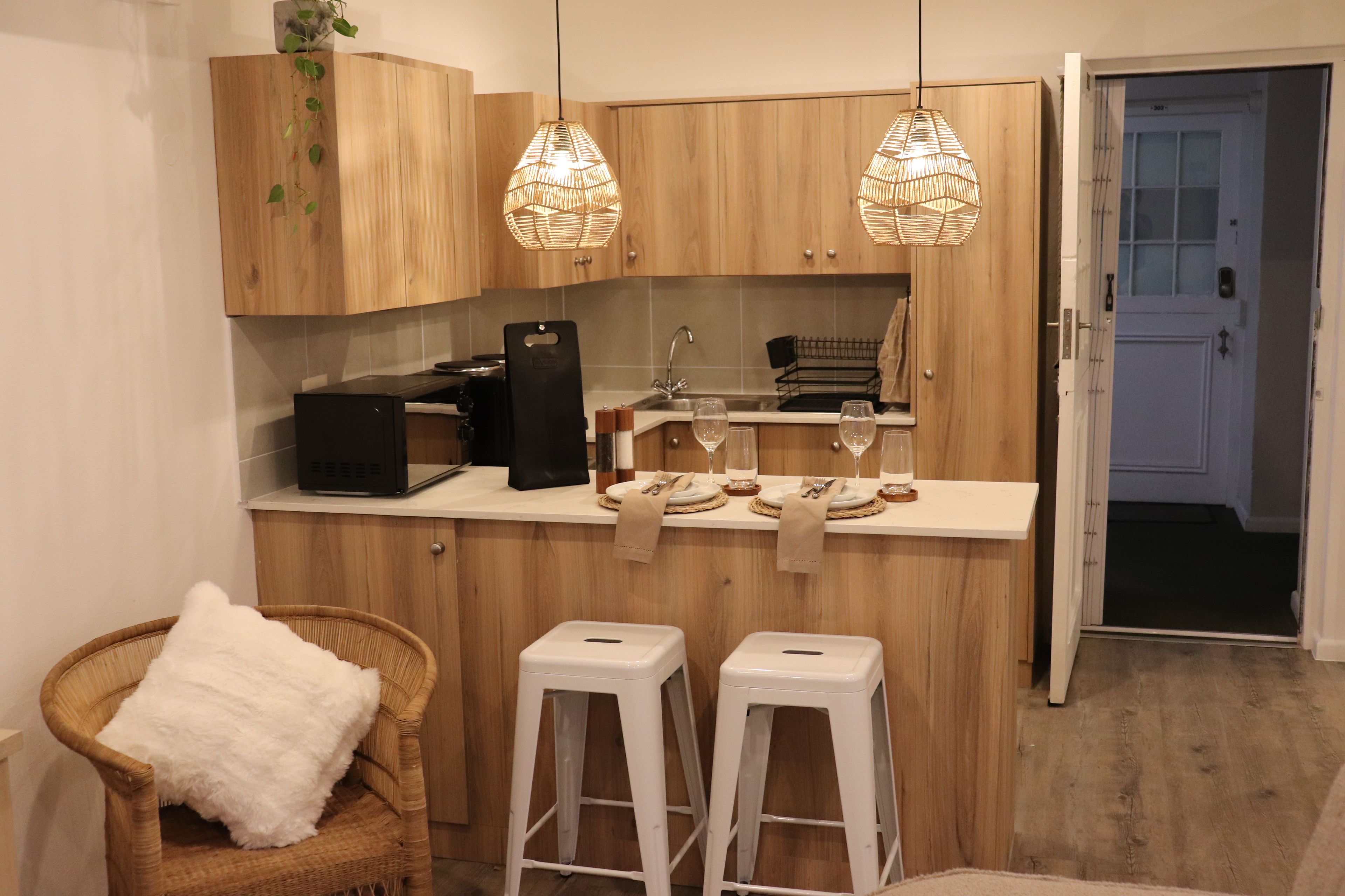 Een kleine keuken met houten kastjes, een zwarte koffieapparaat, twee witte krukken en een rieten stoel met een wit kussen.