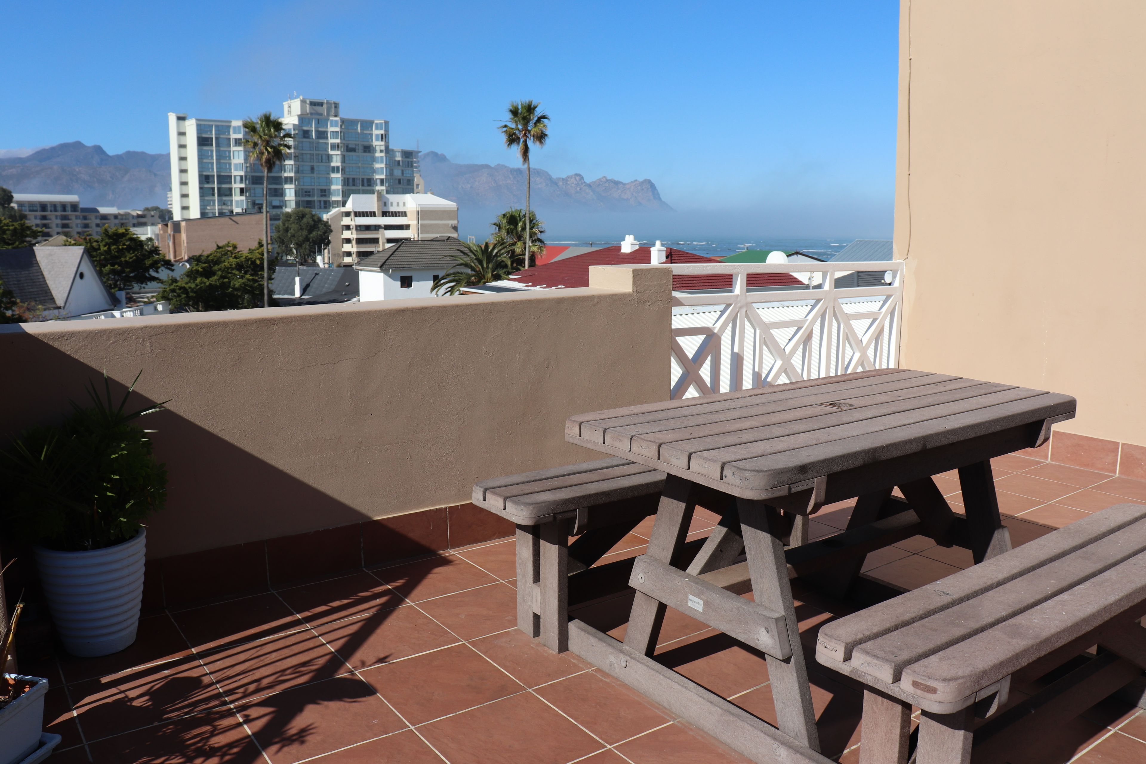 A wooden picnic table with benches on a tiled patio with a view of the ocean and buildings in the background.
