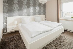 A modern bedroom with a large bed, a nightstand, a lamp, and a window with a view. - Holiday apartment in Norderney with Wi-Fi (Norderney)