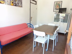 Quarto com sofá vermelho, mesa de madeira, cadeiras brancas e um armário com televisão.