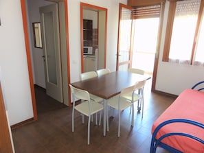 A dining area with a wooden table and four white chairs, a kitchen in the background, and a bed with a red cover to the right. - Family apartment with pool access (Bibione)
