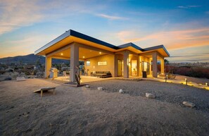 A modern house with a covered patio, outdoor seating, and a desert landscape in the background. - Horizon House | Wellness Retreat Near Joshua Tree (Yucca Valley)