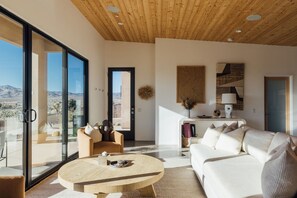 A modern living room with a large glass door leading to a balcony, a round wooden coffee table, a white sofa, and a wooden ceiling. - Horizon House | Wellness Retreat Near Joshua Tree (Yucca Valley)