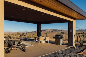 An outdoor patio with a stationary exercise bike, a barbecue grill, and a table with chairs. - Horizon House | Wellness Retreat Near Joshua Tree (Yucca Valley)