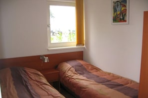 A room with two single beds, a bedside table, a lamp, a window with curtains, and a painting on the wall. - Holiday home with 1 bedroom (Burgerbrug)