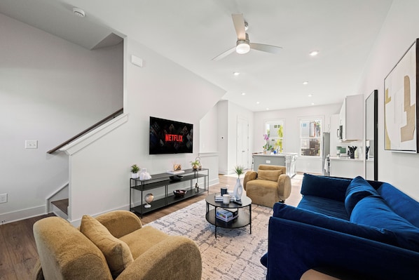 Uma sala de estar moderna com um sofá, poltronas, uma mesa de centro e uma TV exibindo 'NETFLIX'.