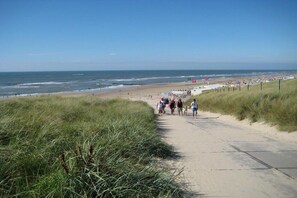 Ein Strandweg, der zum Ufer führt, mit Menschen, die darauf spazieren gehen, einem klaren Himmel und dem Ozean im Hintergrund.