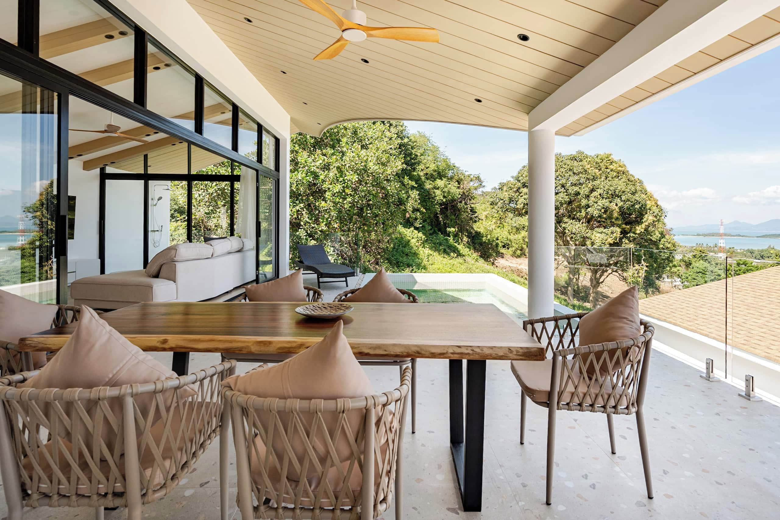 Un espacio moderno para comer al aire libre con una mesa de madera y sillas de mimbre, con vistas a un exuberante jardín y una masa de agua.