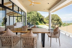 Un espace de restauration extérieur moderne, avec une table en bois et des chaises en osier, offrant une vue sur un jardin luxuriant et un plan d’eau.