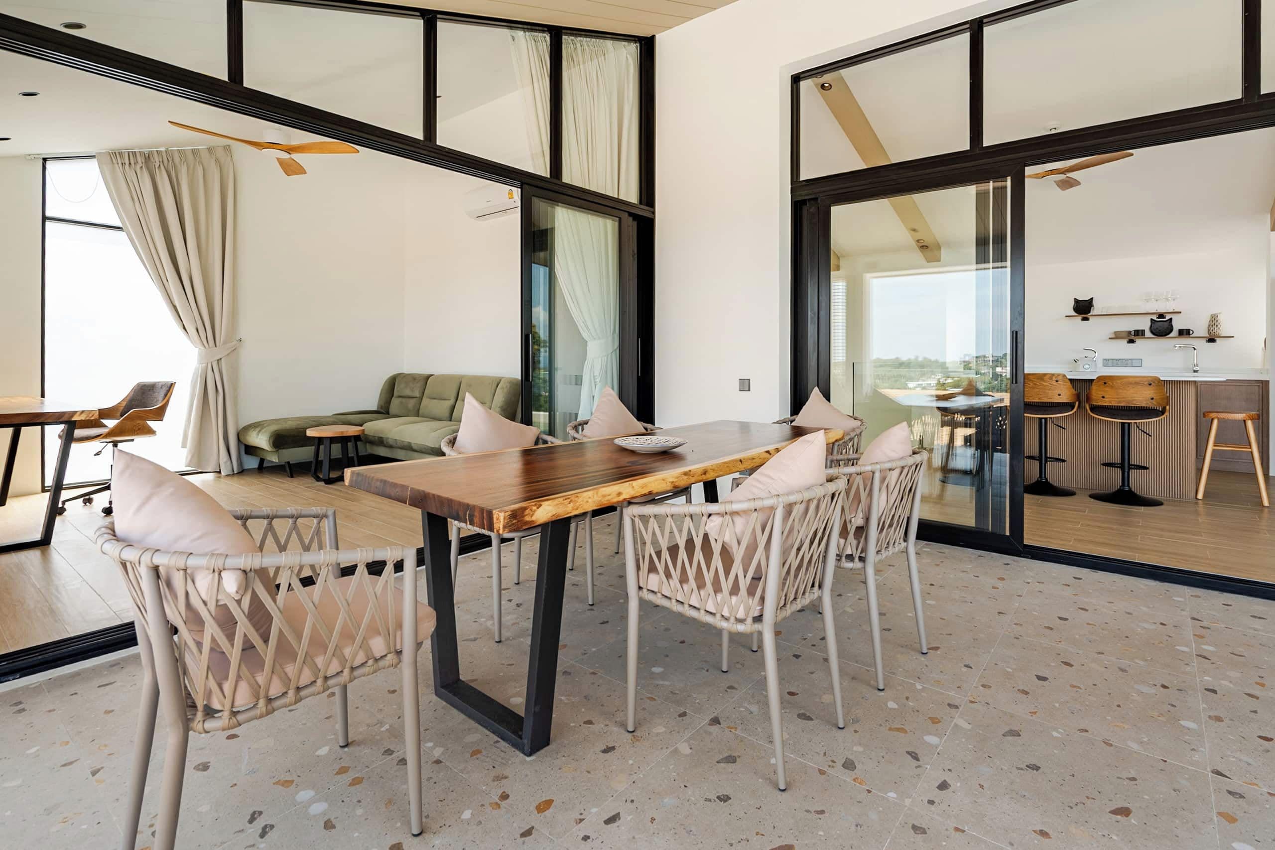 A modern dining area with a wooden table and wicker chairs, a sofa, and a kitchen visible through glass doors.