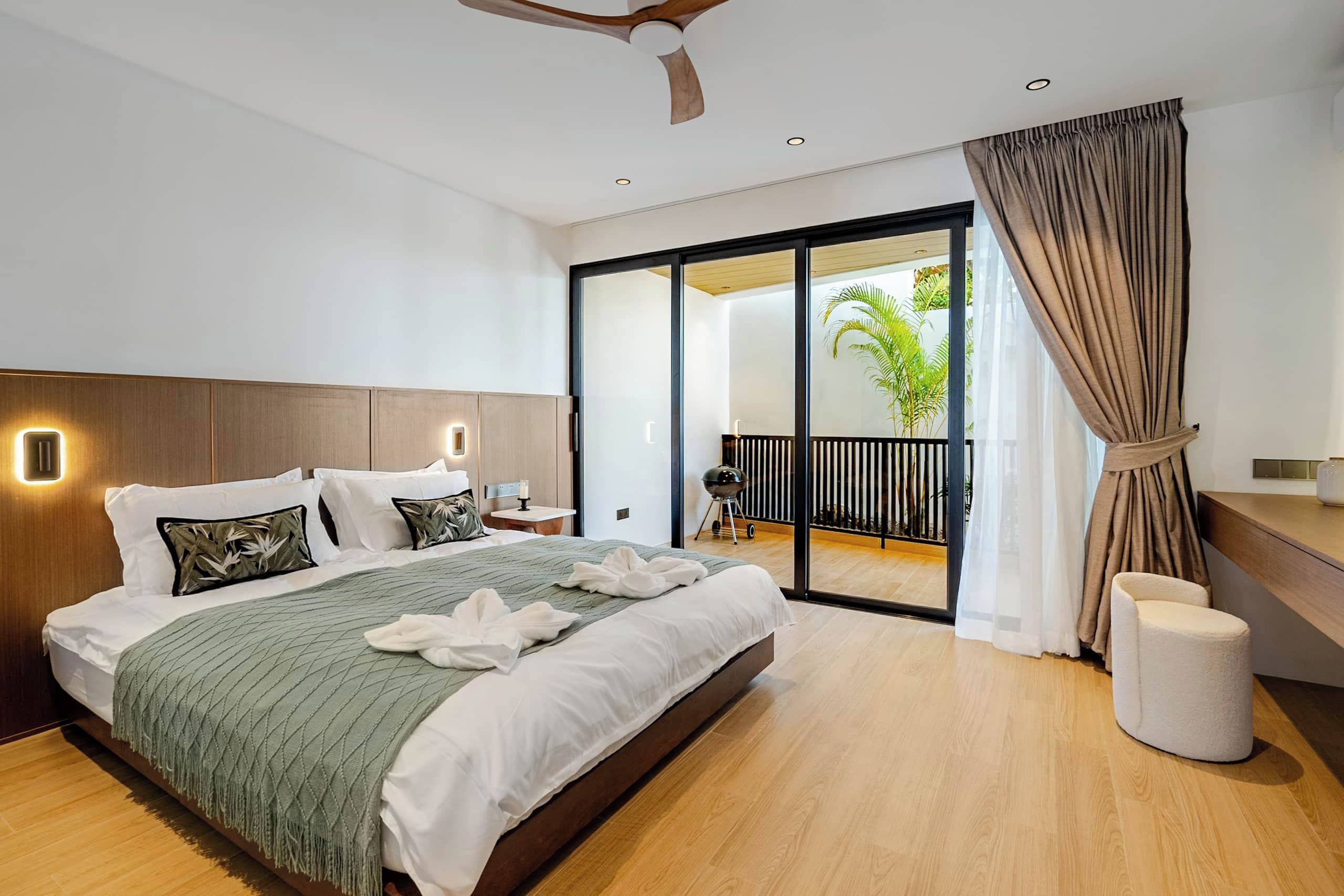 Un dormitorio moderno con una cama grande, un ventilador de techo, un cristalero corredero a un balcón y una mesita de noche pequeña.