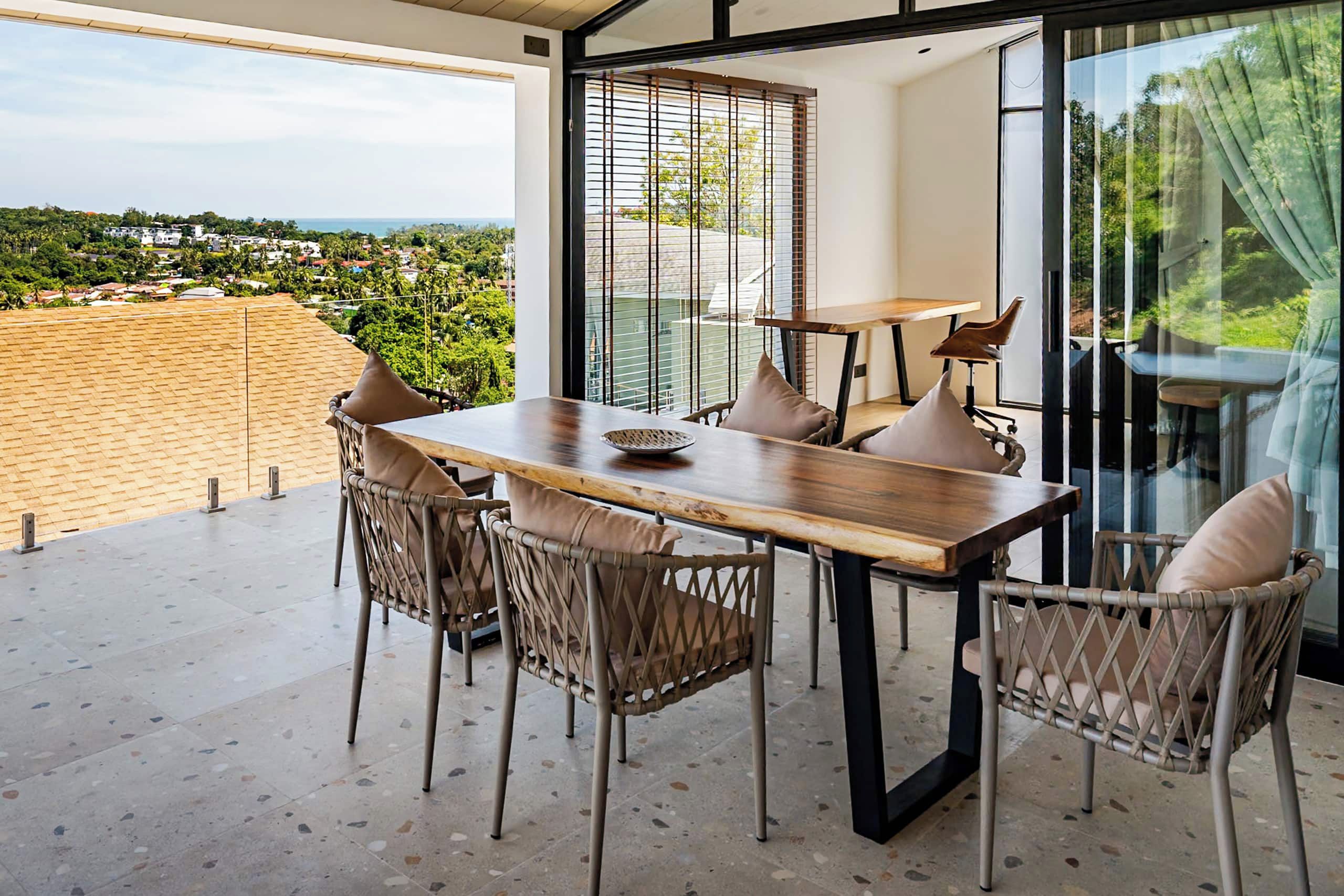 A dining area with a wooden table and wicker chairs, large windows with blinds, and a view of a residential area.