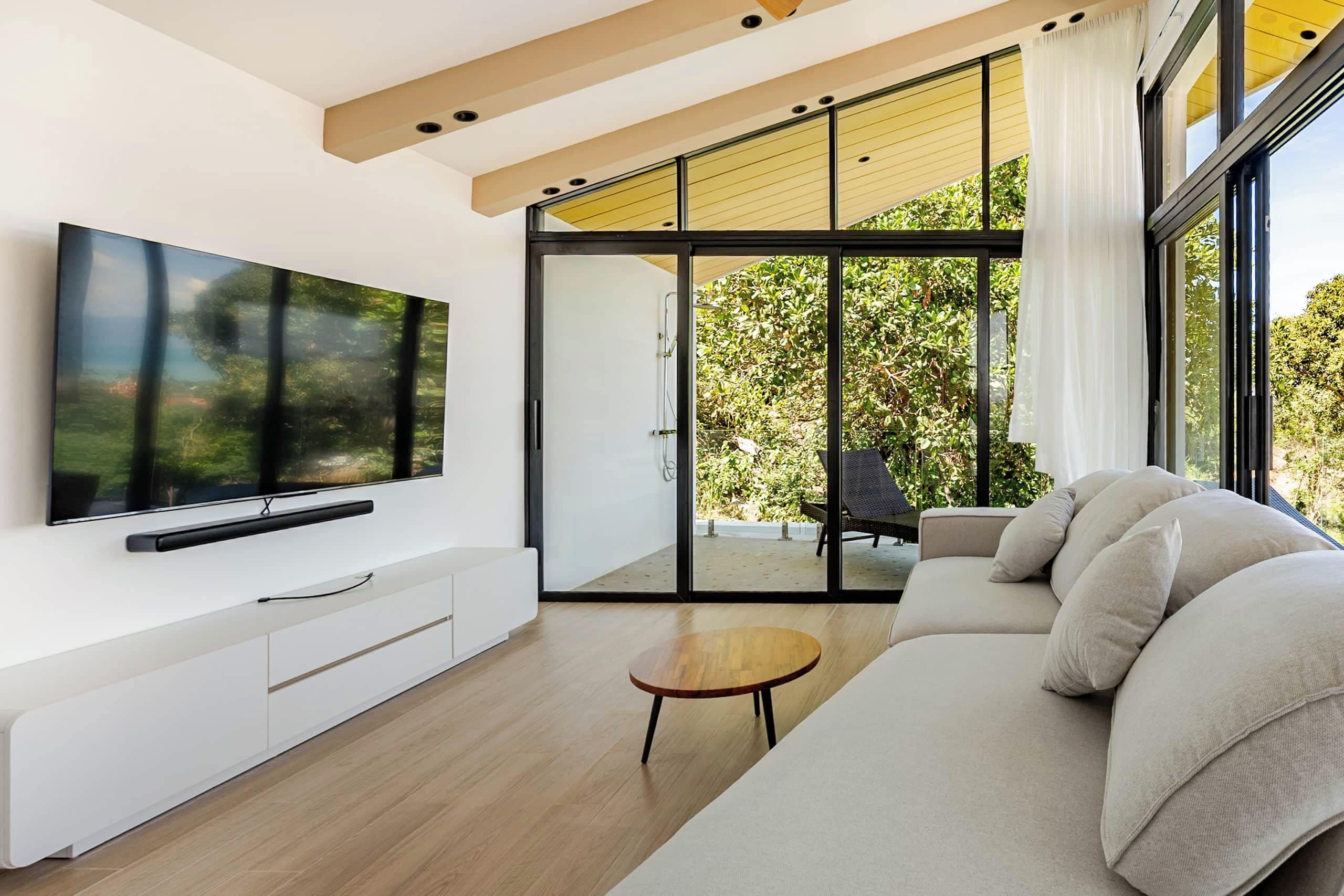 Uma sala de estar moderna com uma TV de tela plana, um sofá, uma pequena mesa de madeira e uma porta de vidro deslizante que dá para uma área externa.