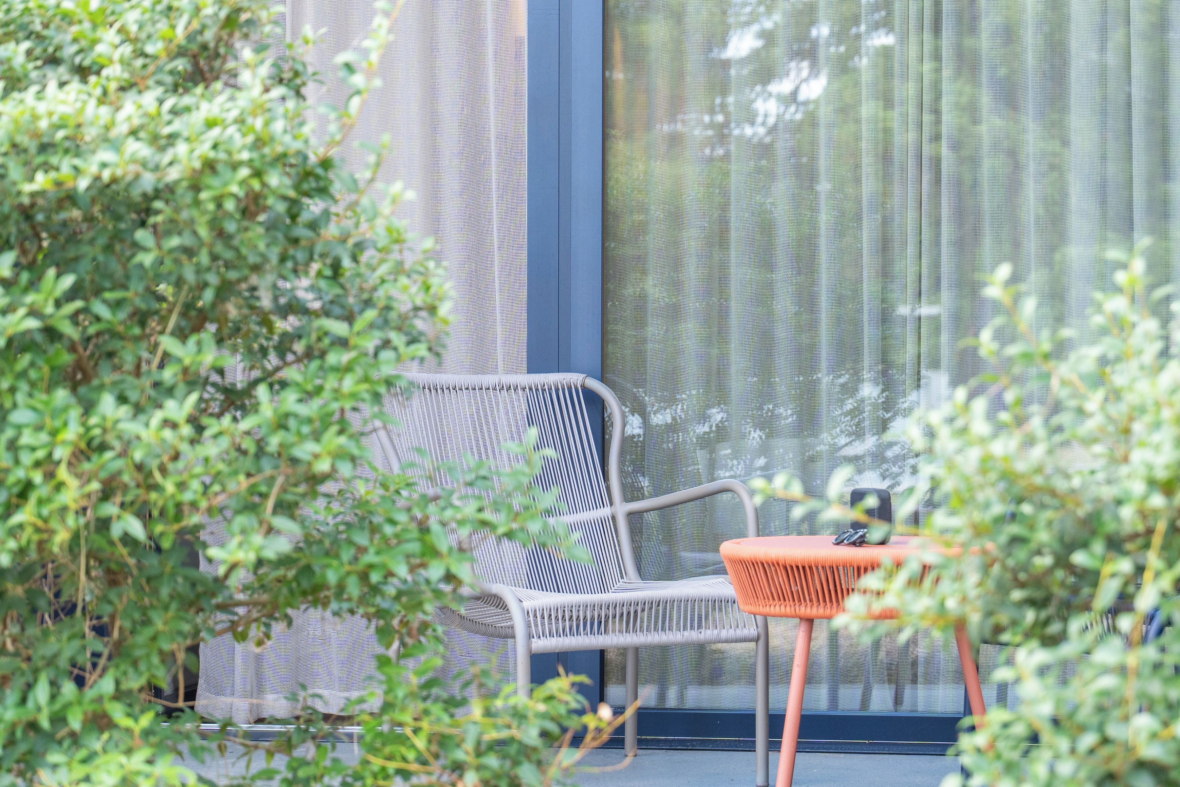 Una silla de mimbre y una mesita con cojín, ubicadas frente a una ventana con cortina estampada.