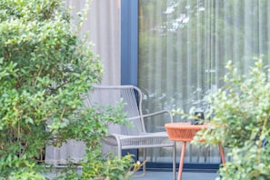 Una silla de mimbre y una mesita con cojín, ubicadas frente a una ventana con cortina estampada.