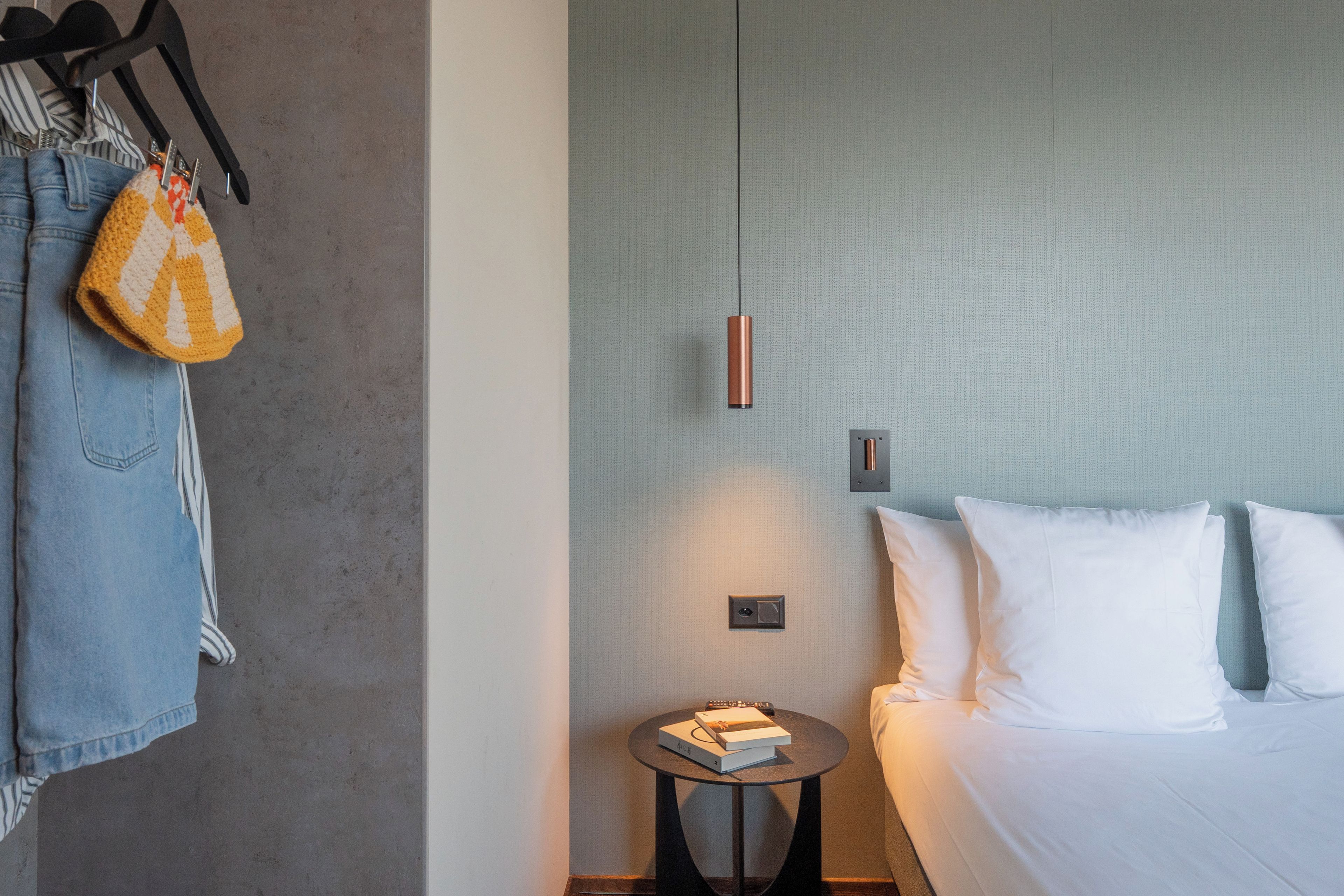 Ein Hotelzimmer mit einem Bett, einem Nachttisch mit einem Buch, einer Pendelleuchte und einem Kleiderständer mit Jacke und Tasche.