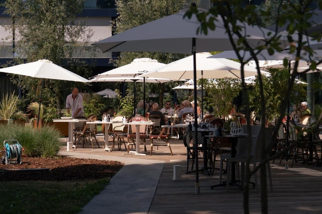 Un área de comedor al aire libre con mesas y sillas bajo sombrillas, rodeada de follaje.