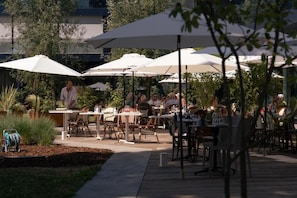 An outdoor dining area with tables and chairs under umbrellas, surrounded by greenery. - Pattùs Hôtel (LA GRANDE BÉROCHE)