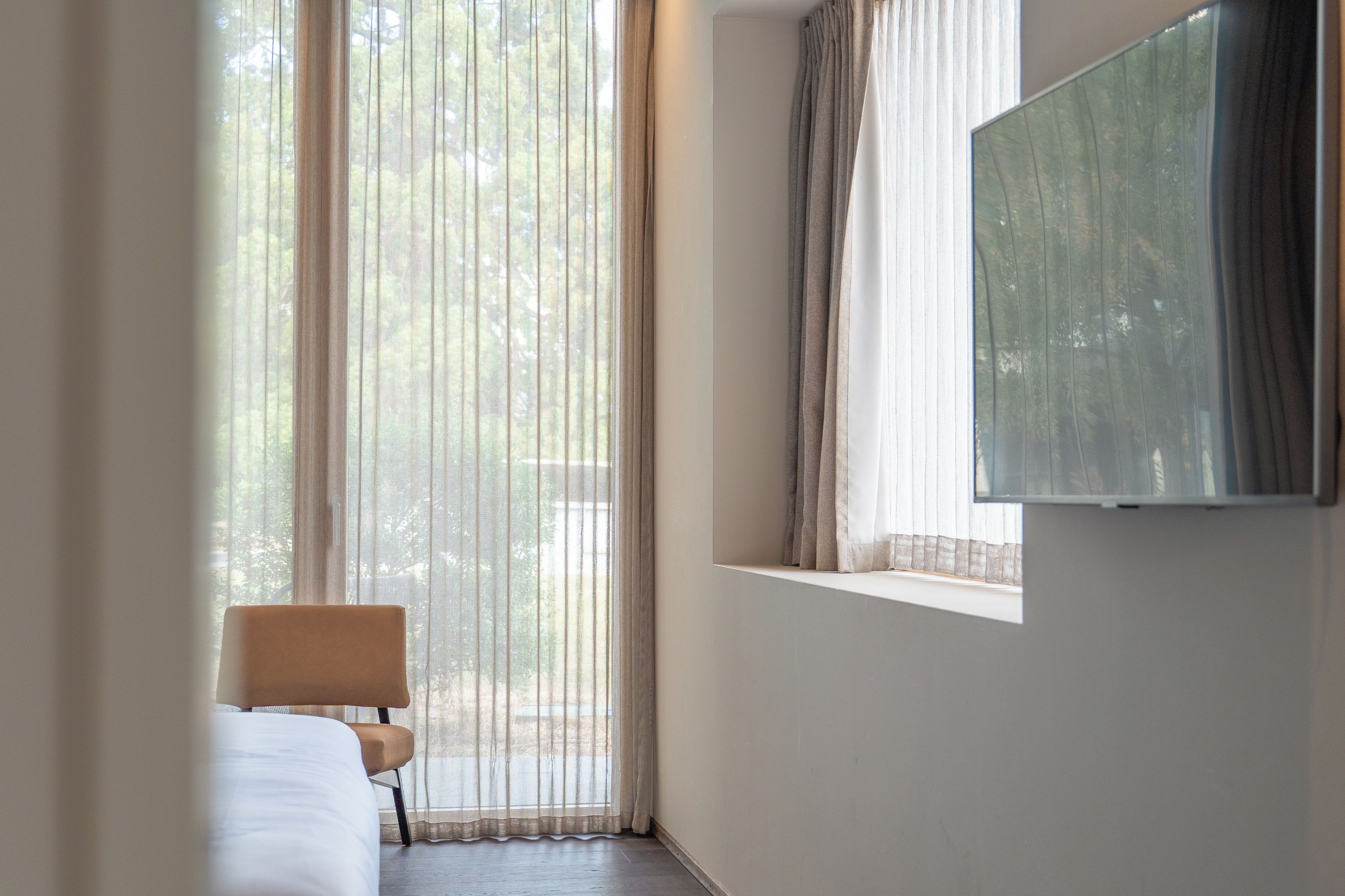 Habitación de hotel con cama, una silla, televisión montada en la pared y ventana con cortinas traslúcidas.
