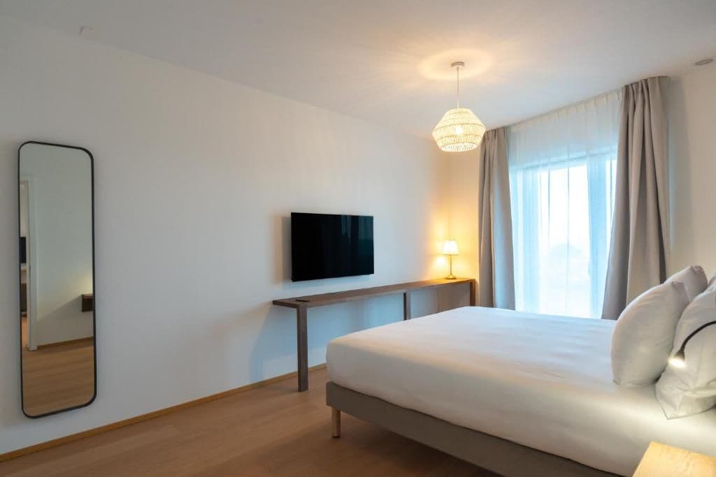 A hotel room with a bed, a flat-screen TV mounted on the wall, a bedside table with a lamp, and a large window with curtains.