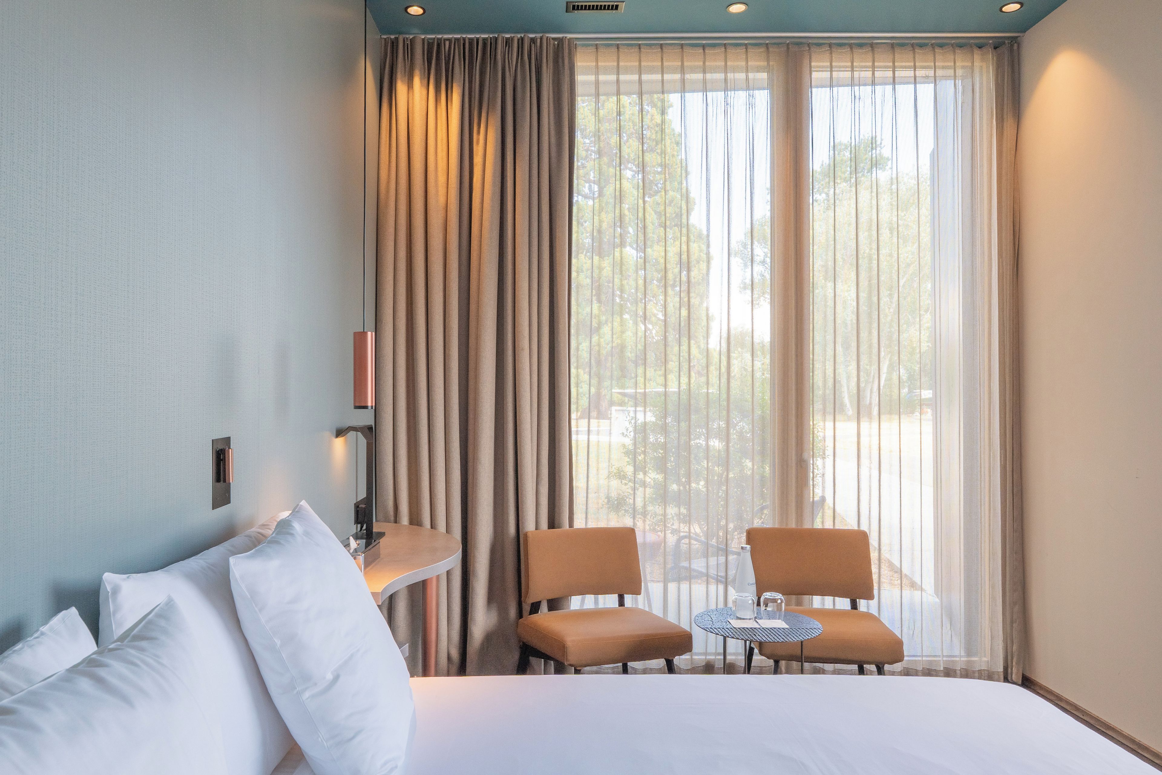 Una habitación de hotel con cama, dos sillas, una mesita y un ventanal con cortinas.