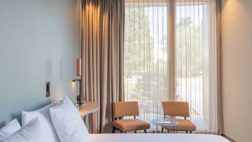 Una habitación de hotel con cama, dos sillas, una mesita y un ventanal con cortinas.