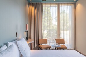 Une chambre d’hôtel avec un lit, deux chaises, une petite table et une fenêtre avec des rideaux.