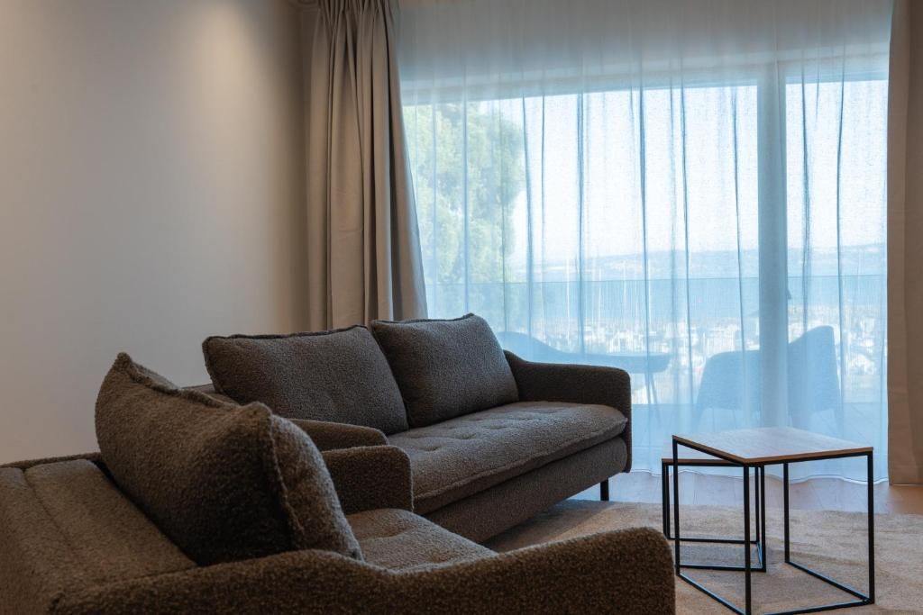 Una sala de estar moderna con un sofá y una mesita, ventanas amplias con cortinas translúcidas, y vistas al exterior.