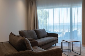 A modern living room with a sofa and a small table, large windows with sheer curtains, and a view of the outdoors. - Pattùs Hôtel (La Grande-Beroche)