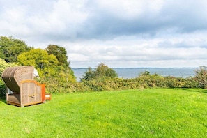 A grassy area with a wicker chair and a small table, overlooking a body of water and trees. - Holiday home with 2 bedrooms (Langballig)