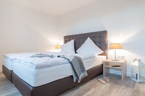 A neatly made bed with pillows and a blanket, two bedside tables with lamps, and a pair of slippers on the floor. - Holiday apartment with 2 bedrooms (Börgerende)