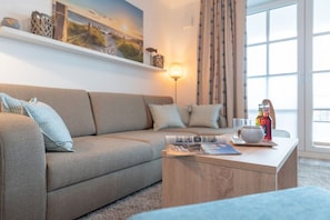 A modern living room with a sofa, a coffee table with magazines, a lamp, and a window with curtains. - Holiday apartment with 2 bedrooms (Börgerende)