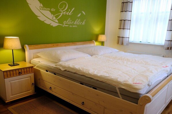 Ein Zimmer mit einem Einzelbett, einem Holzrahmen, einer grünen Wand mit einer Feder und Text, zwei Nachttischlampen und einem mit Vorhängen versehenen Fenster.