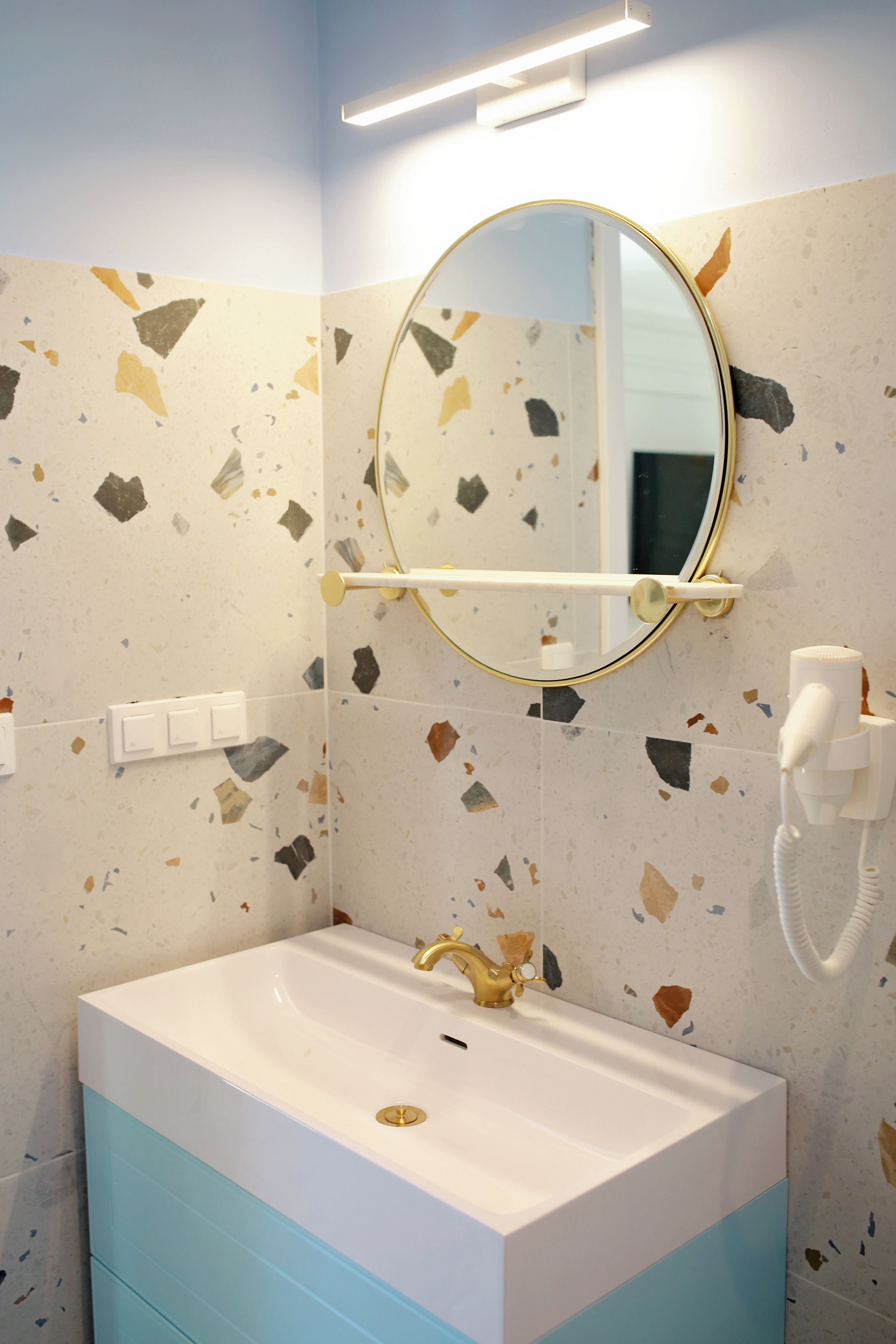 A bathroom with a white sink, gold faucet, and a round mirror. There is a telephone on the wall and a light fixture above the mirror.