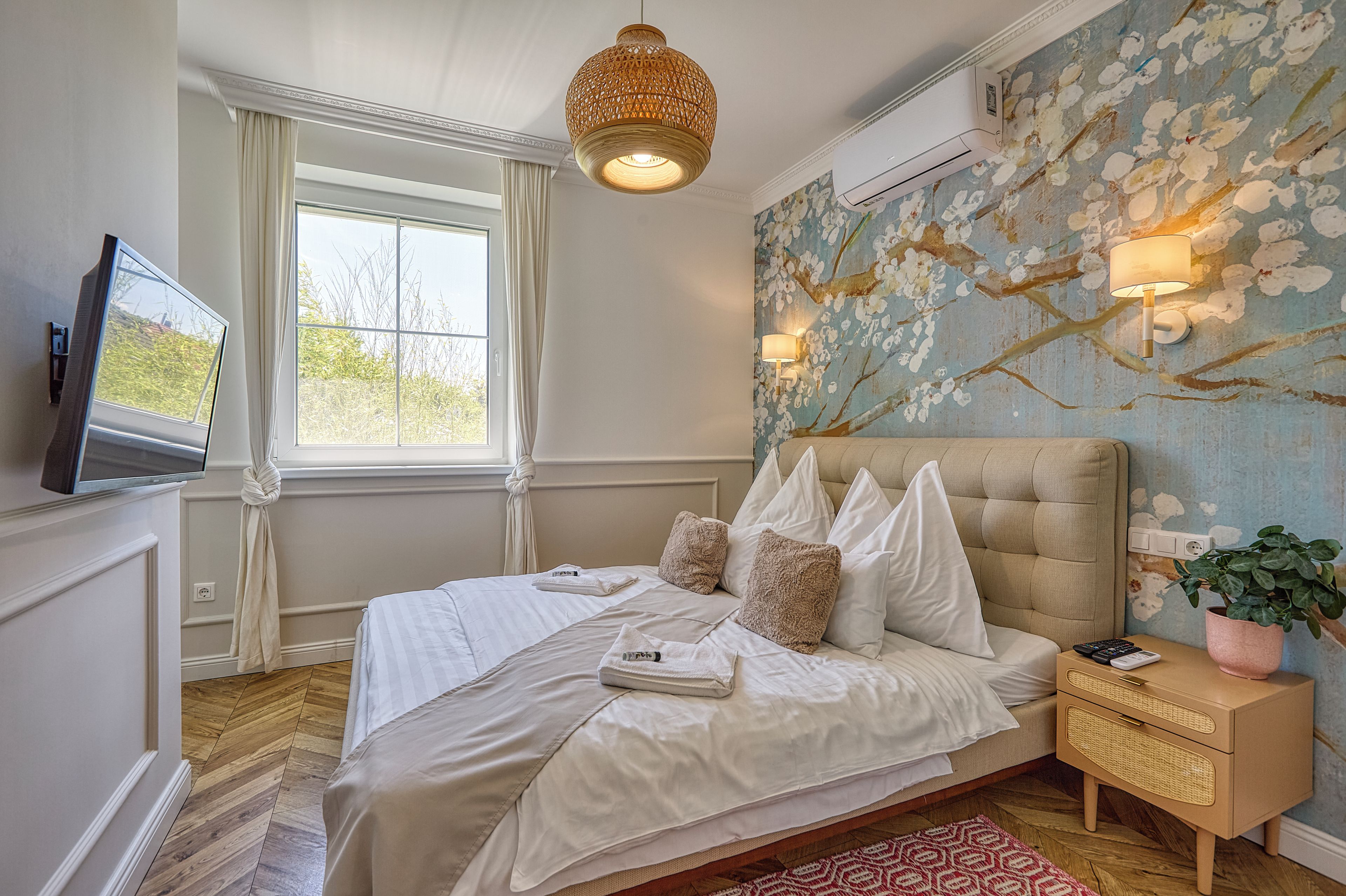 Ein Schlafzimmer mit einem Bett, einem Fernseher, einem Fenster, einer Wandlampe und einem Nachttisch mit einer Pflanze.
