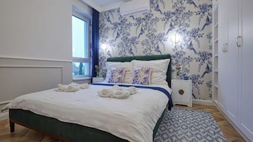 Una cama bien tendida con sábanas blancas y almohadones decorativos, un papel tapiz floral azul y blanco, y un pequeño aparador blanco con tirador de latón.