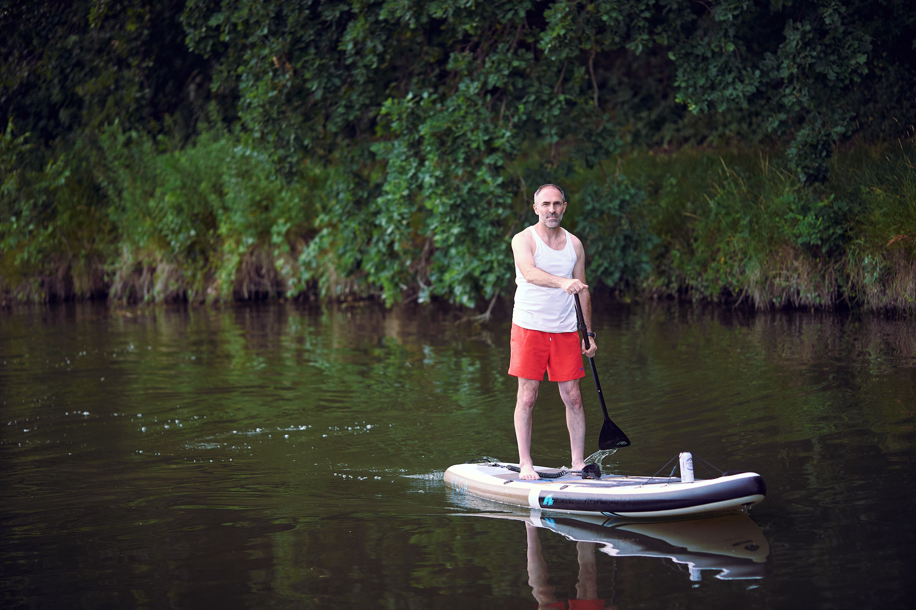 Une personne se tient debout sur une planche de paddle, sur une étendue d’eau, avec une végétation luxuriante en arrière-plan.