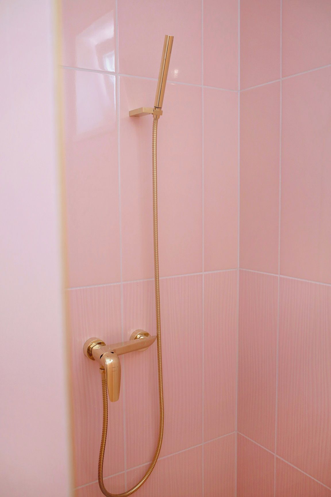 Chuveiro com chuveiro e mangueira dourados em frente a uma parede de azulejos cor-de-rosa.