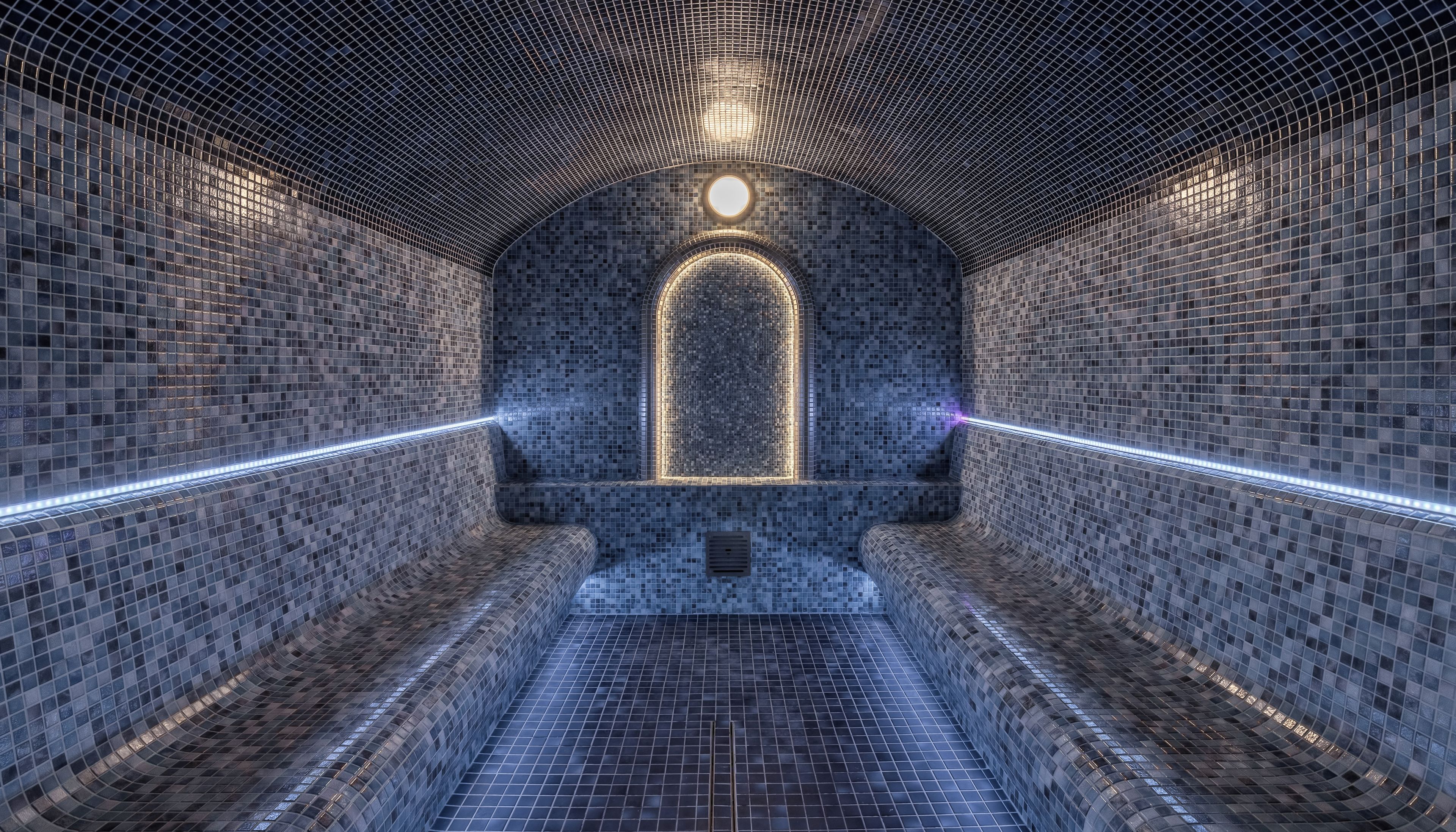 Une structure en forme de tunnel, dotée d’un plafond et de murs ornés de motifs, d’une arche centrale illuminée et d’un sol à motifs géométriques.