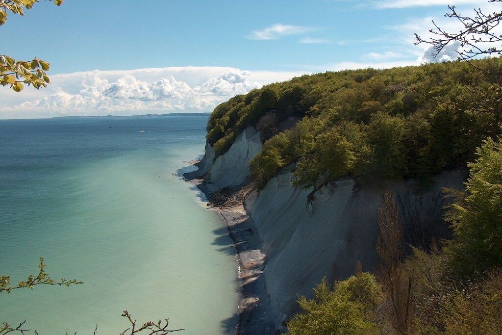 A coastal landscape with a cliff overlooking a calm sea, surrounded by dense greenery.