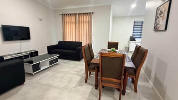 A modern living room with a flat-screen TV, a black sofa, a wooden dining table with chairs, and a large window with curtains.