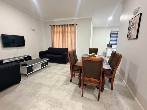 A modern living room with a flat-screen TV, a black sofa, a wooden dining table with chairs, and a large window with curtains. - Dreams Apartment 115 (Victoria Falls)
