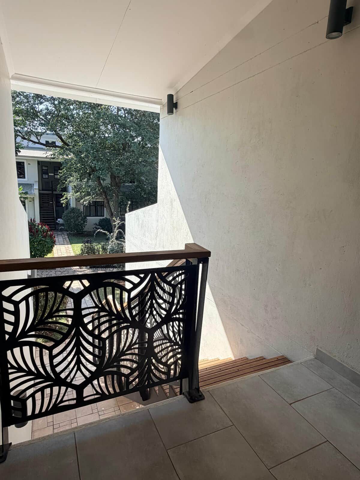 A balcony with a geometric metal railing, a tiled floor, and a view of a residential area with trees and a building.
