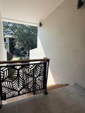 A balcony with a geometric metal railing, a tiled floor, and a view of a residential area with trees and a building. - Dreams Apartment 115 (Victoria Falls)