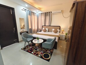 A hotel room with a bed, two armchairs, a small table, a bedside table with a lamp, and an air conditioning unit. - hotel roomin (Noida)