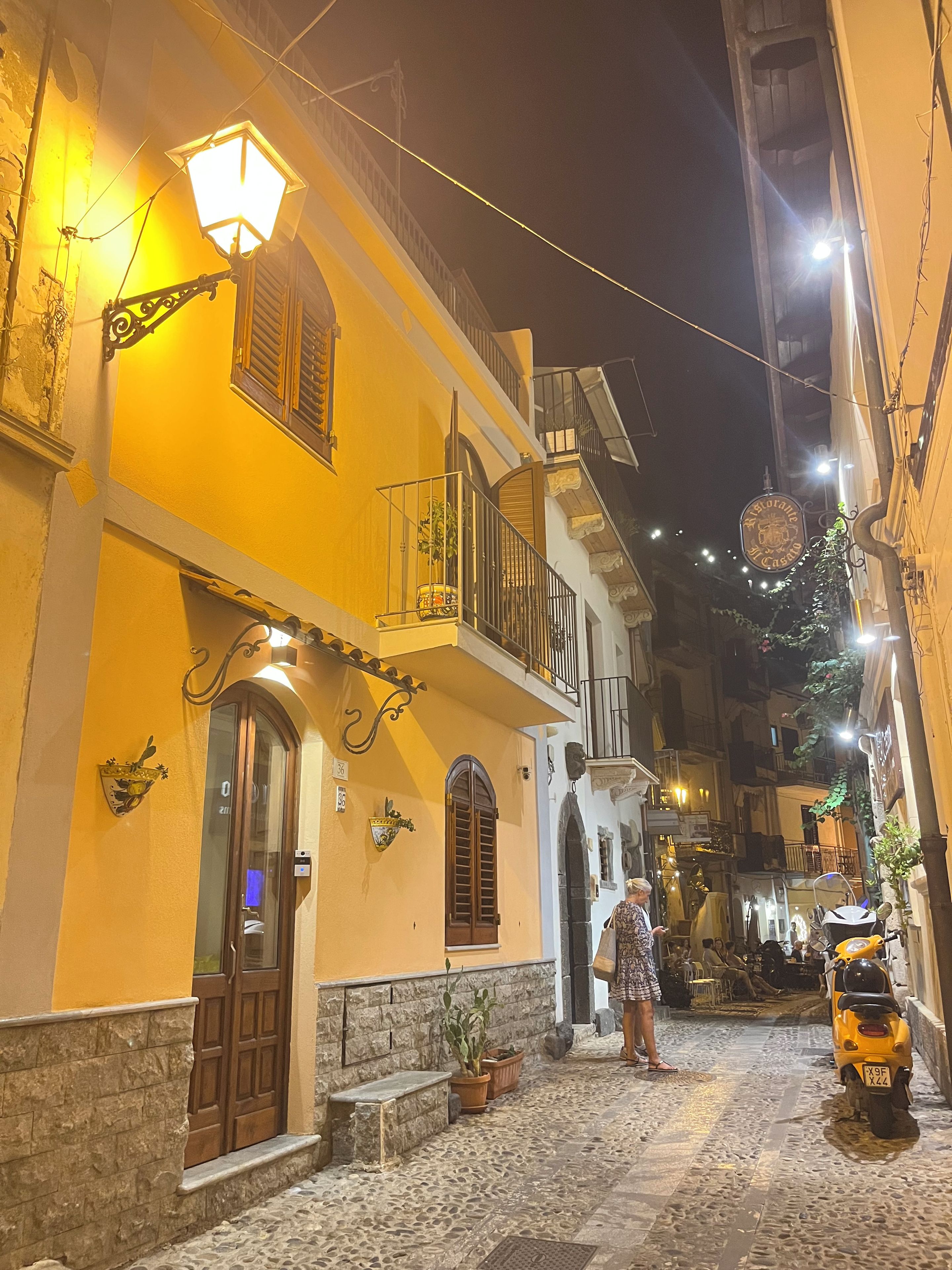 Una stretta via acciottolata con un edificio giallo che presenta un balcone con ringhiera, una porta e una finestra con persiane.